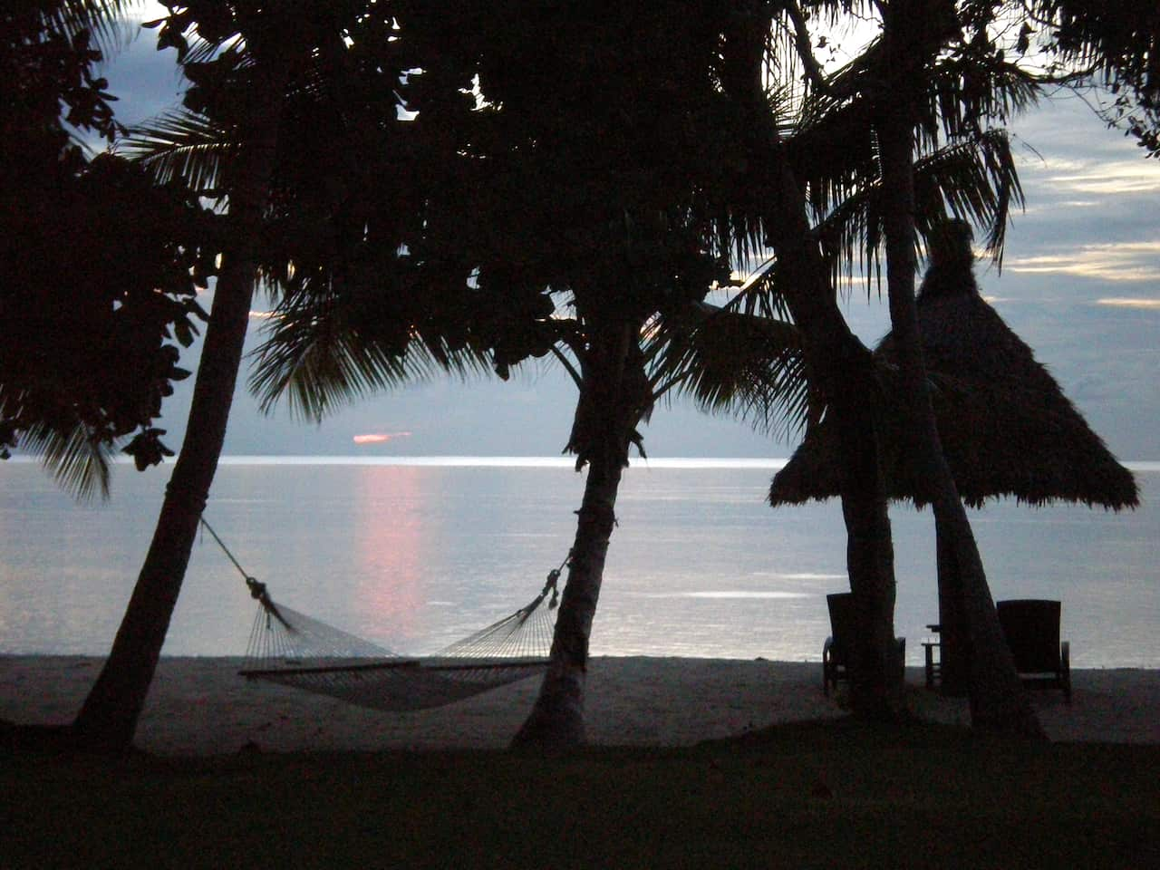 A hammock hangs between two trees, with a beach and sunset in the background.