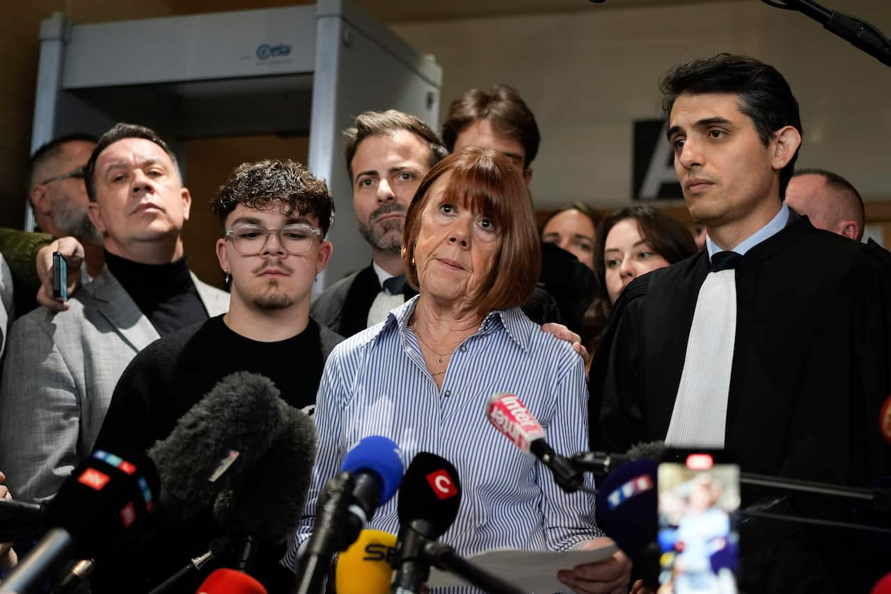 A woman standing behind a row of press microphones with a group of supporters around her.