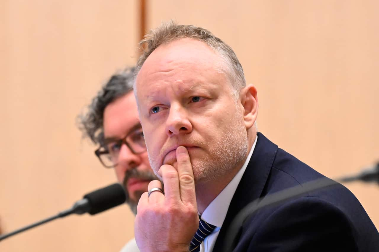 A man wearing a dark suit looks thoughtfully to his left while cradling his chin in his hands. 