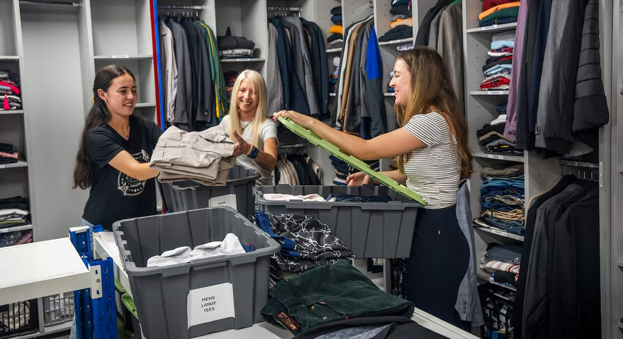 Three women are placing clothes into plastic boxes while standing in a room with wooden shelves full of men's clothing on either side.