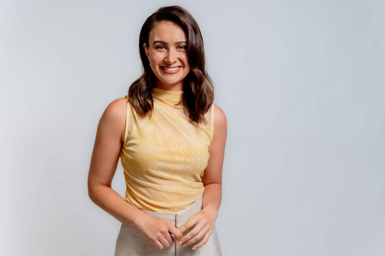 A woman with brown hair and wearing a yellow shirt stands and smiles into the camera