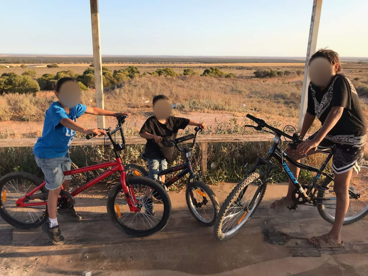 Three young Indigenous boys sitting on bicycles on the verandah of an outback property.