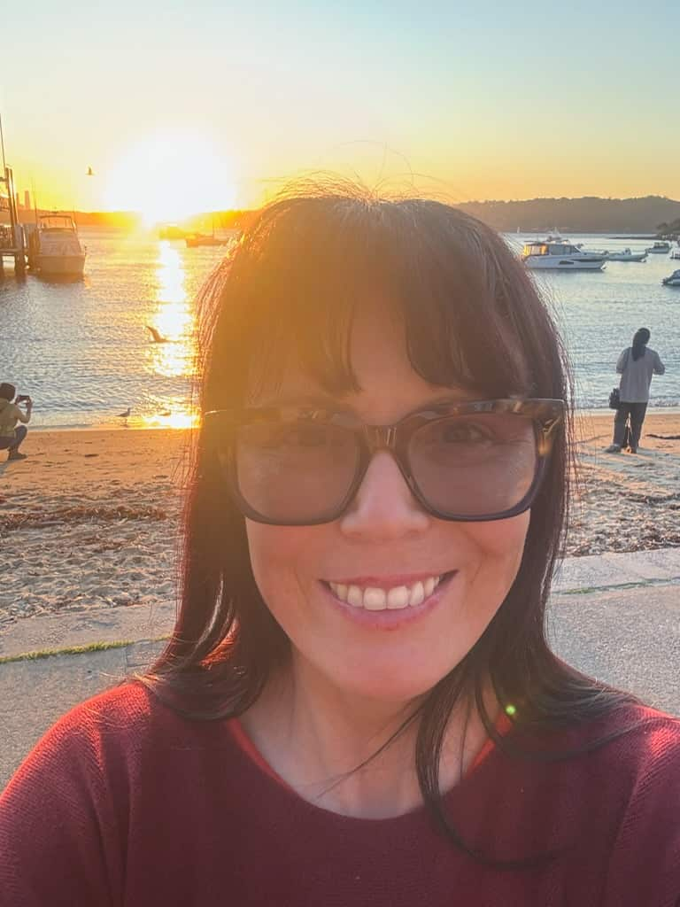 A selfie of a woman at the beach. She is wearing sunglasses and is smiling.