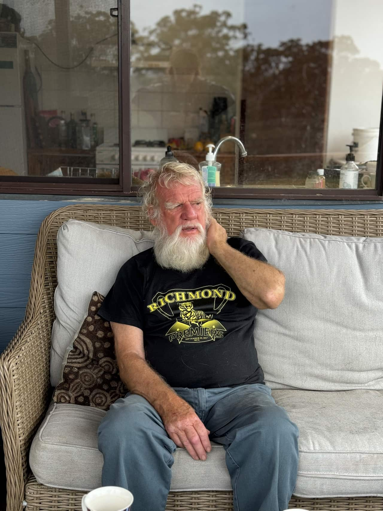 An elderly man with a white beard, wearing a black top and blue jeans, is sitting on a couch outside with one hand on his neck.