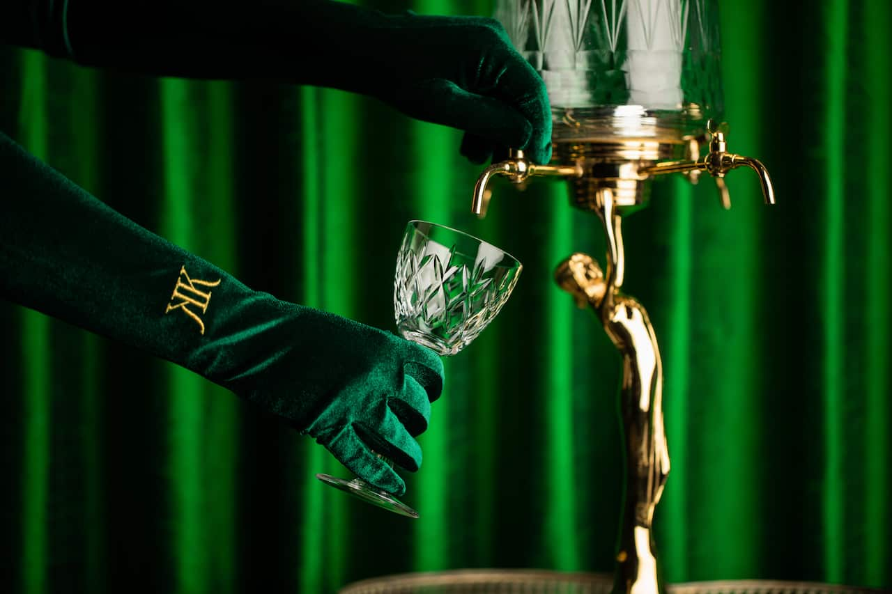 A person wearing green gloves pours a drink into a crystal goblet in front of a green backdrop.