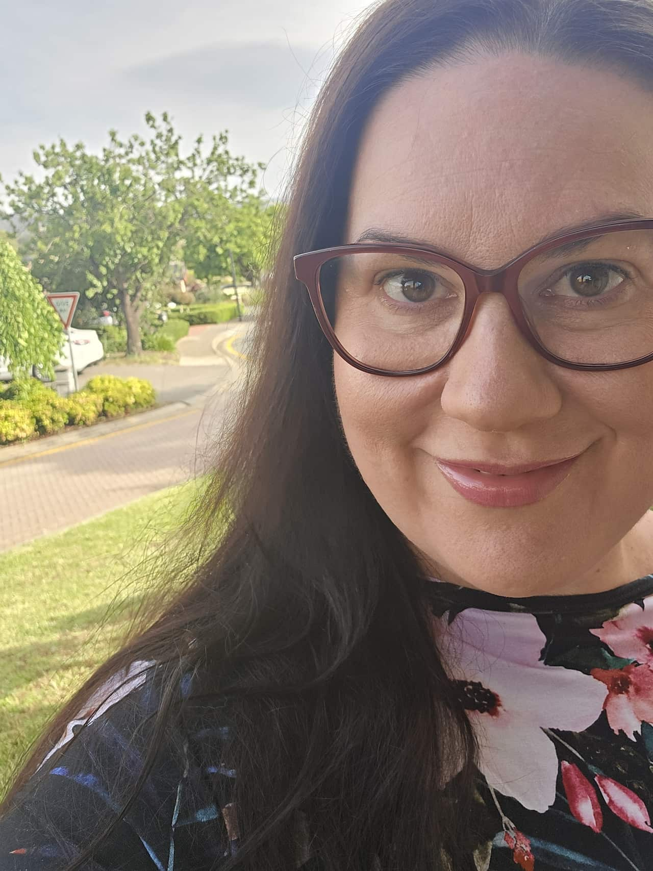 A woman with glasses and long, brown hair wearing a floral dress smiles on a leafy residential street