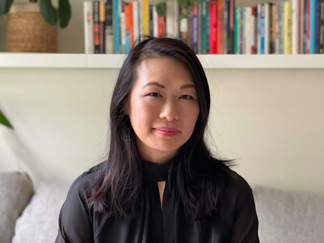 A woman in a black top sits on a grey couch in front of a bookshelf filled with books and a plant in a pot