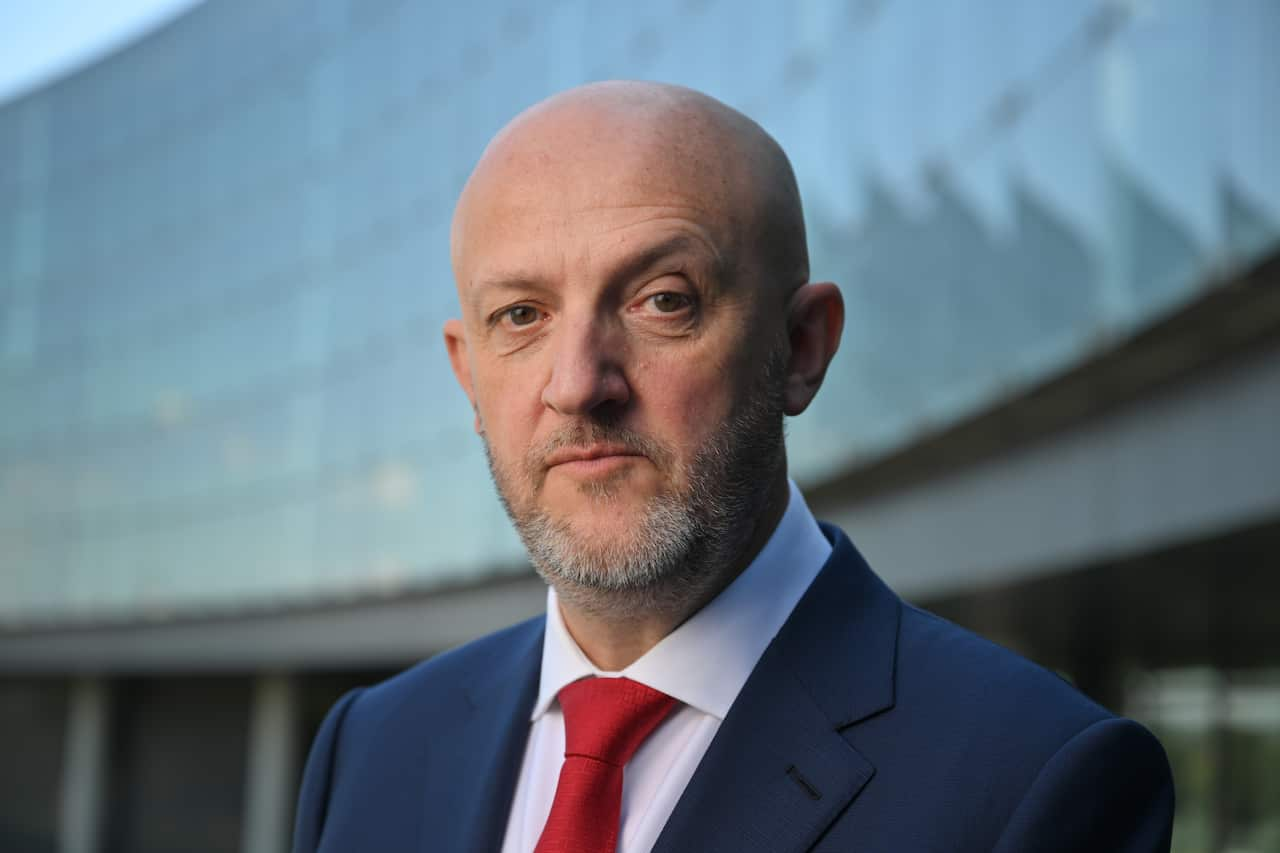 A bald man in a blue suit and a red tie looks at the camera.