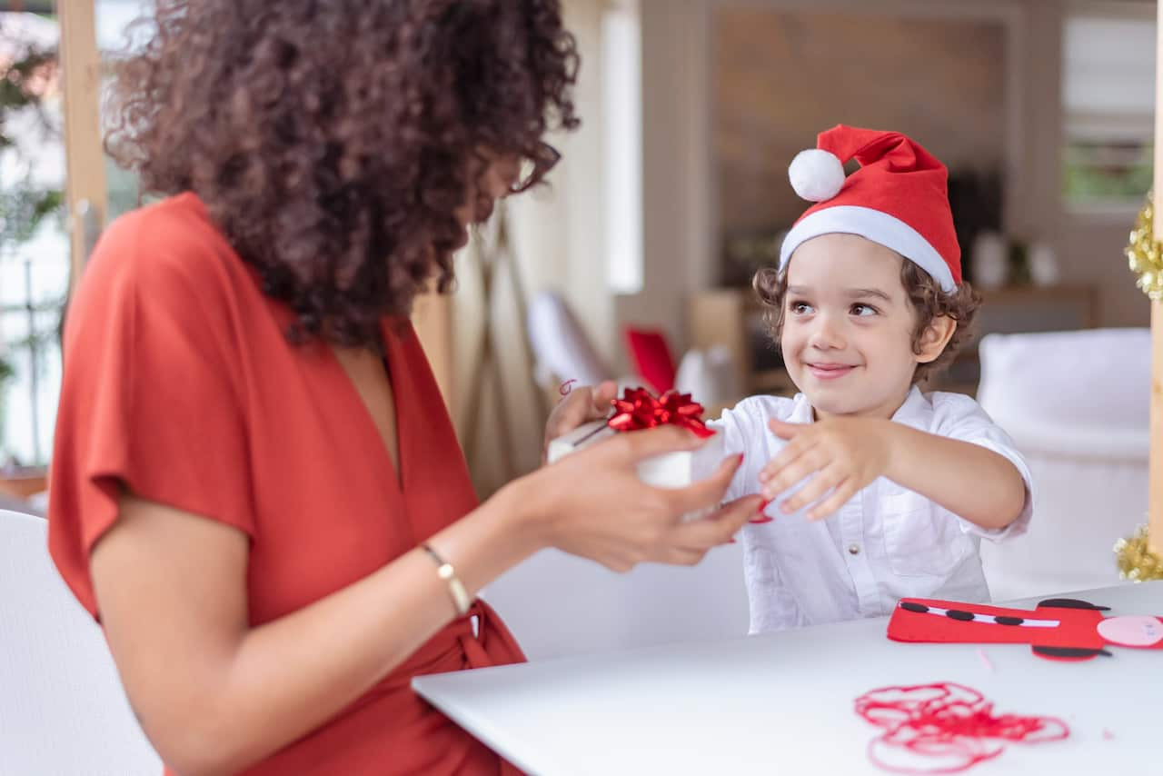 A boy wearing a Santa hat sitting at a table next to a woman who is receiving a small white box with a red bow from him