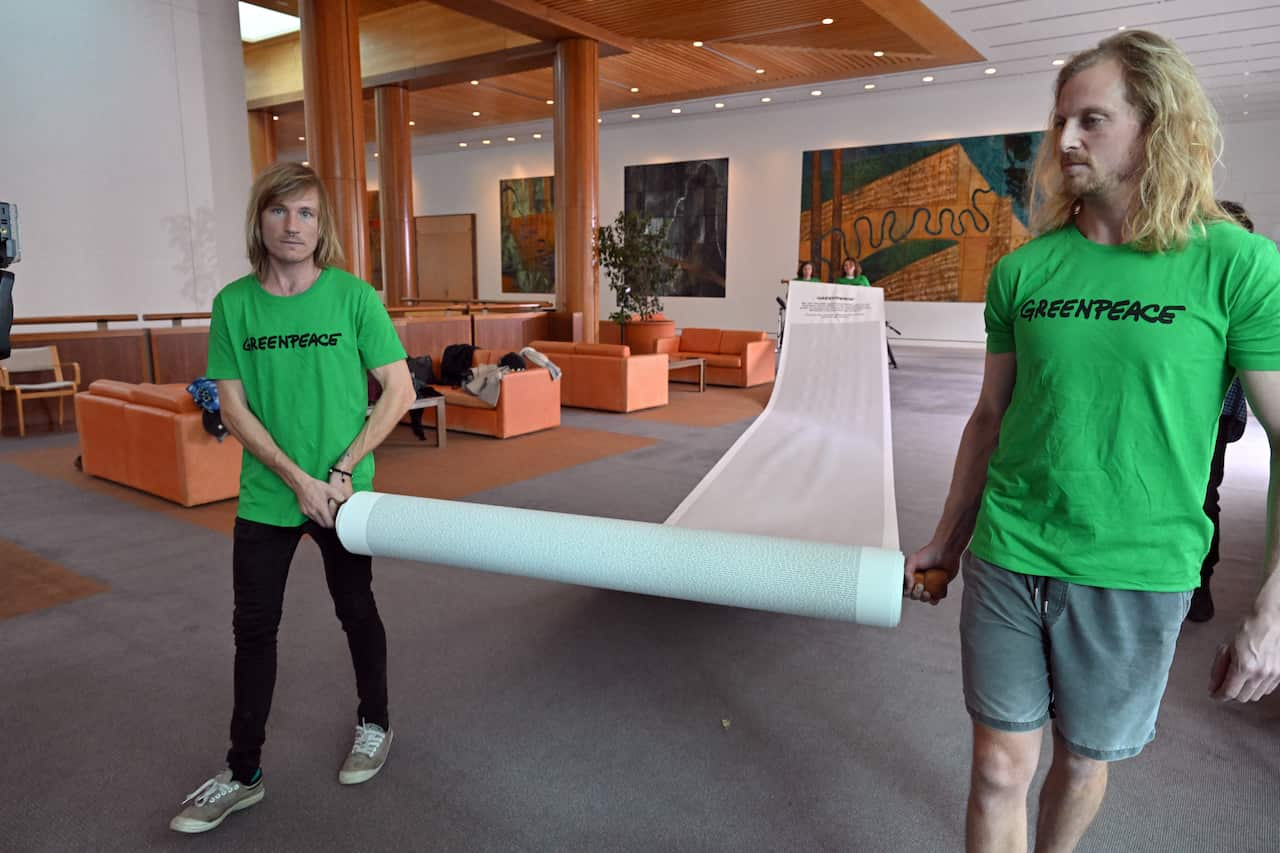 Two men with long hair, wearing green crew-neck t-shirts, unroll a large paper roll as they walk.