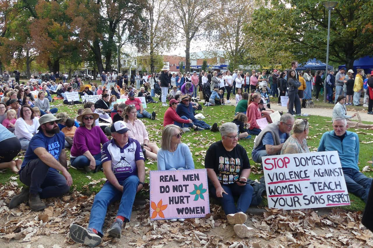 A crowd at a rally against violence towards women, with some sitting and others standing, many holding posters.