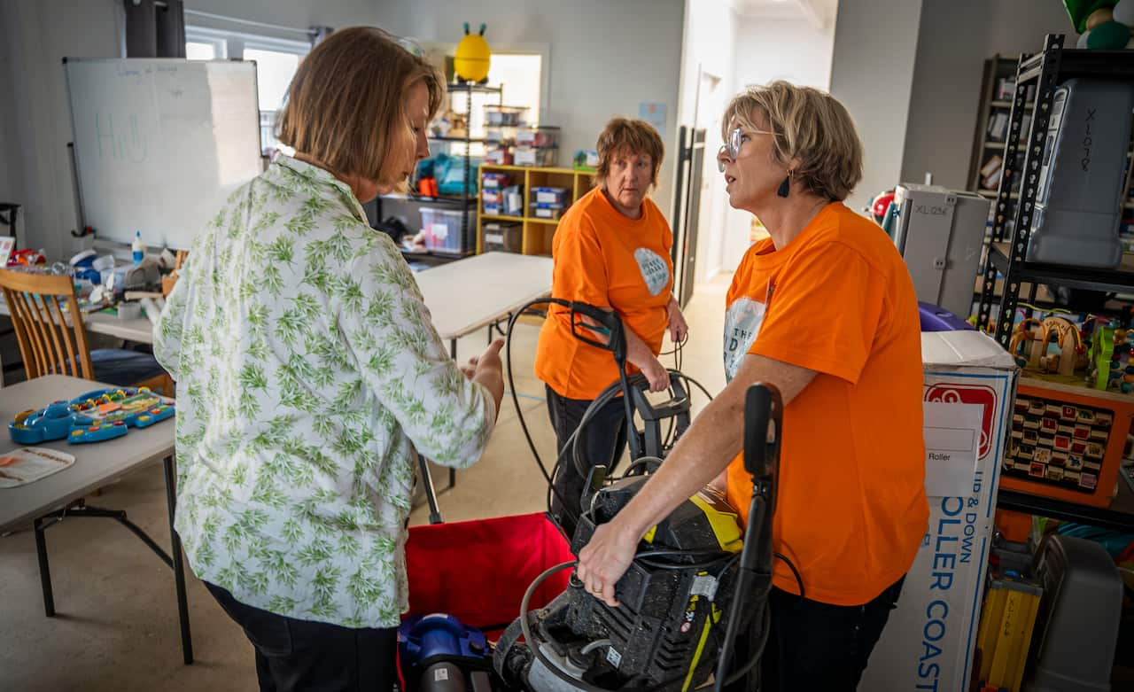 Three women stand in a cluttered room around a trolley with garden tools inside it. 