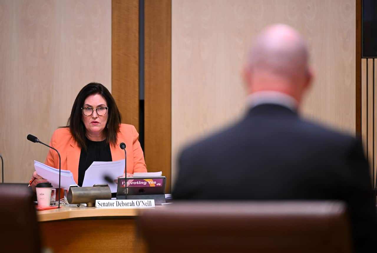 A brunette woman wearing a peach jacket and black t-shirt speaks to a man in a suit during a parliamentary inquiry.