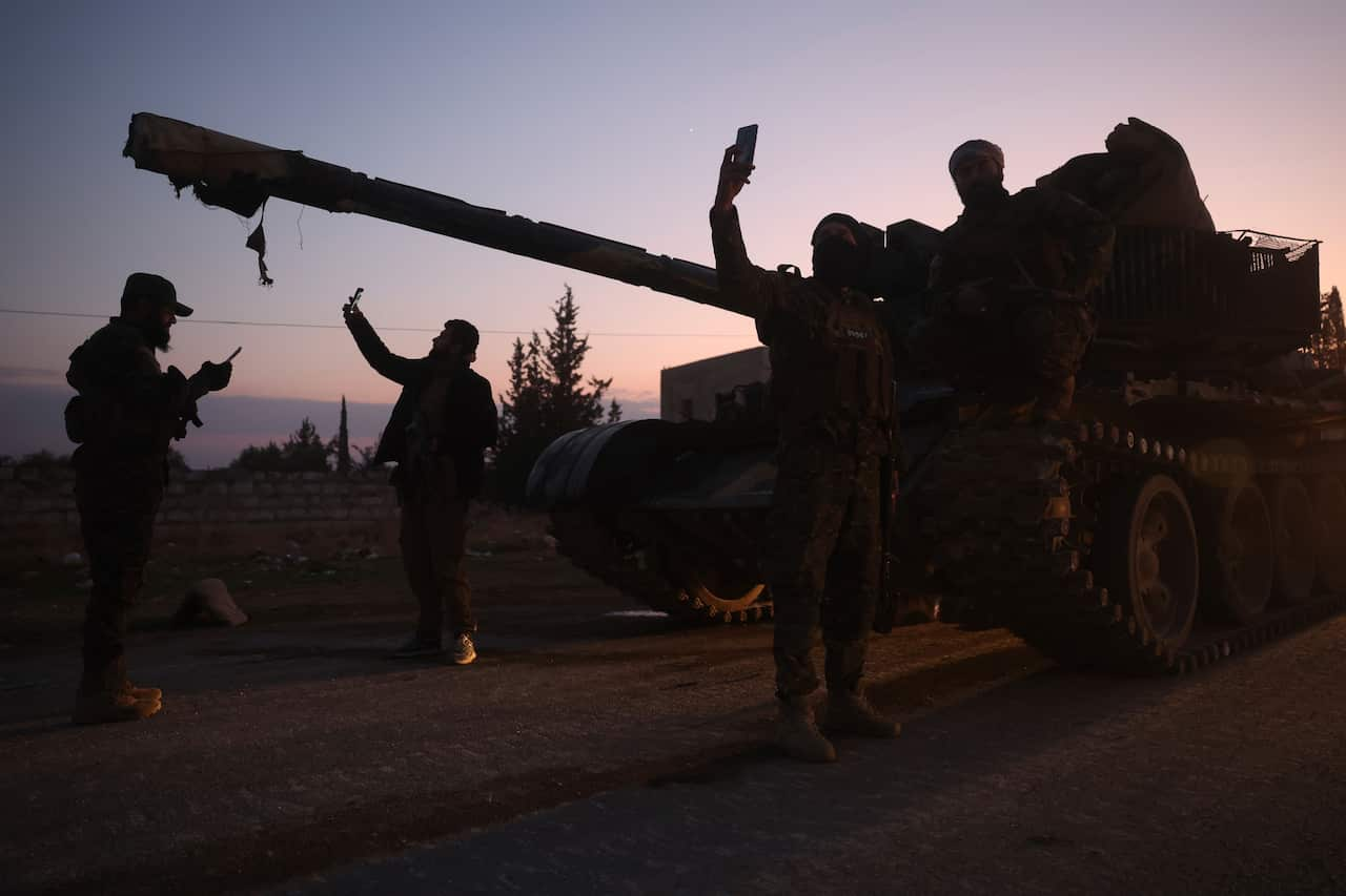 Silhouetted people stand near a tank on a road. Some are taking selfies.