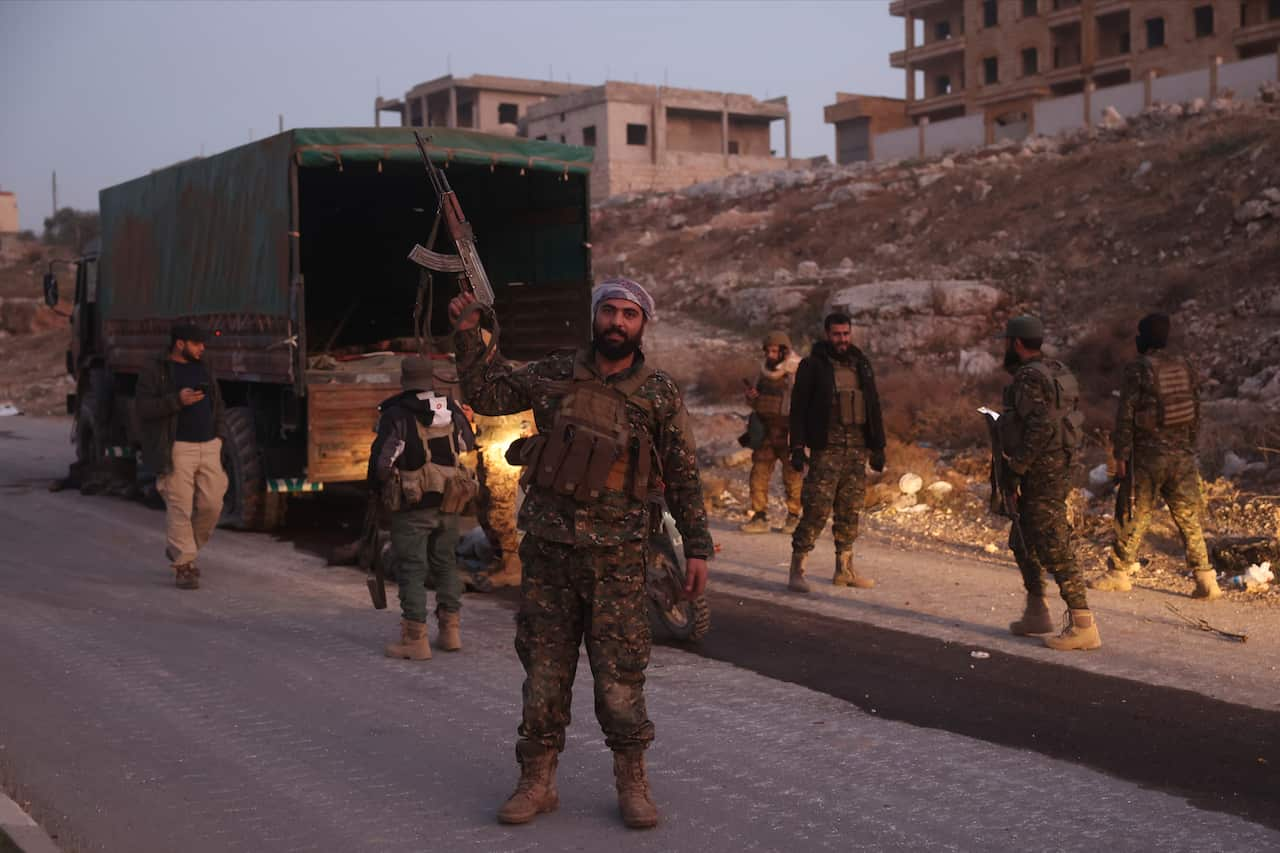 Several men wearing military fatigues stand around a truck. One holds a rifle up in the air.