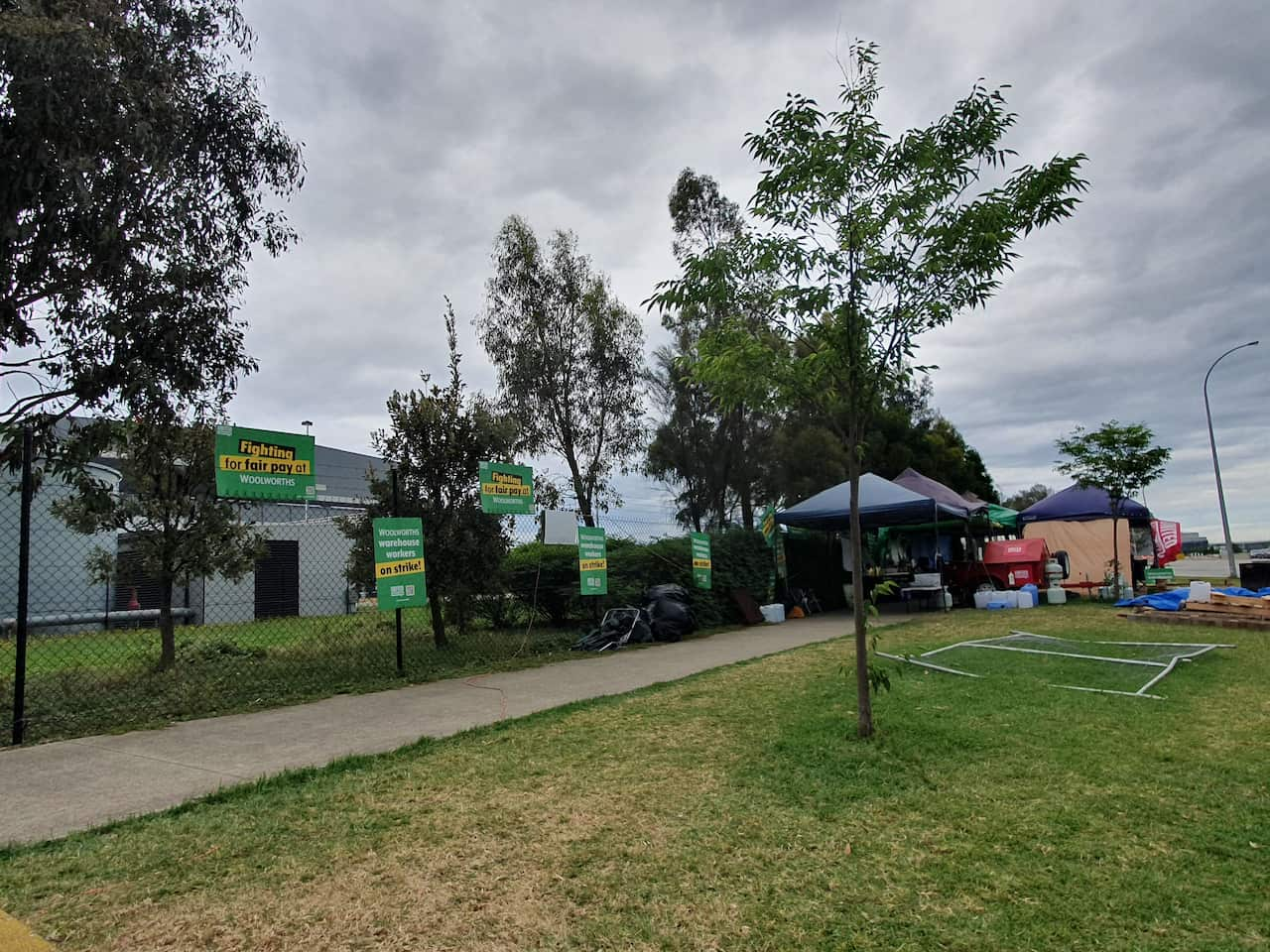 Several blue marquees are set up outside a warehouse fence. Posted to the fence are several signs with slogans such as "Fighting for fair pay at Woolworths".