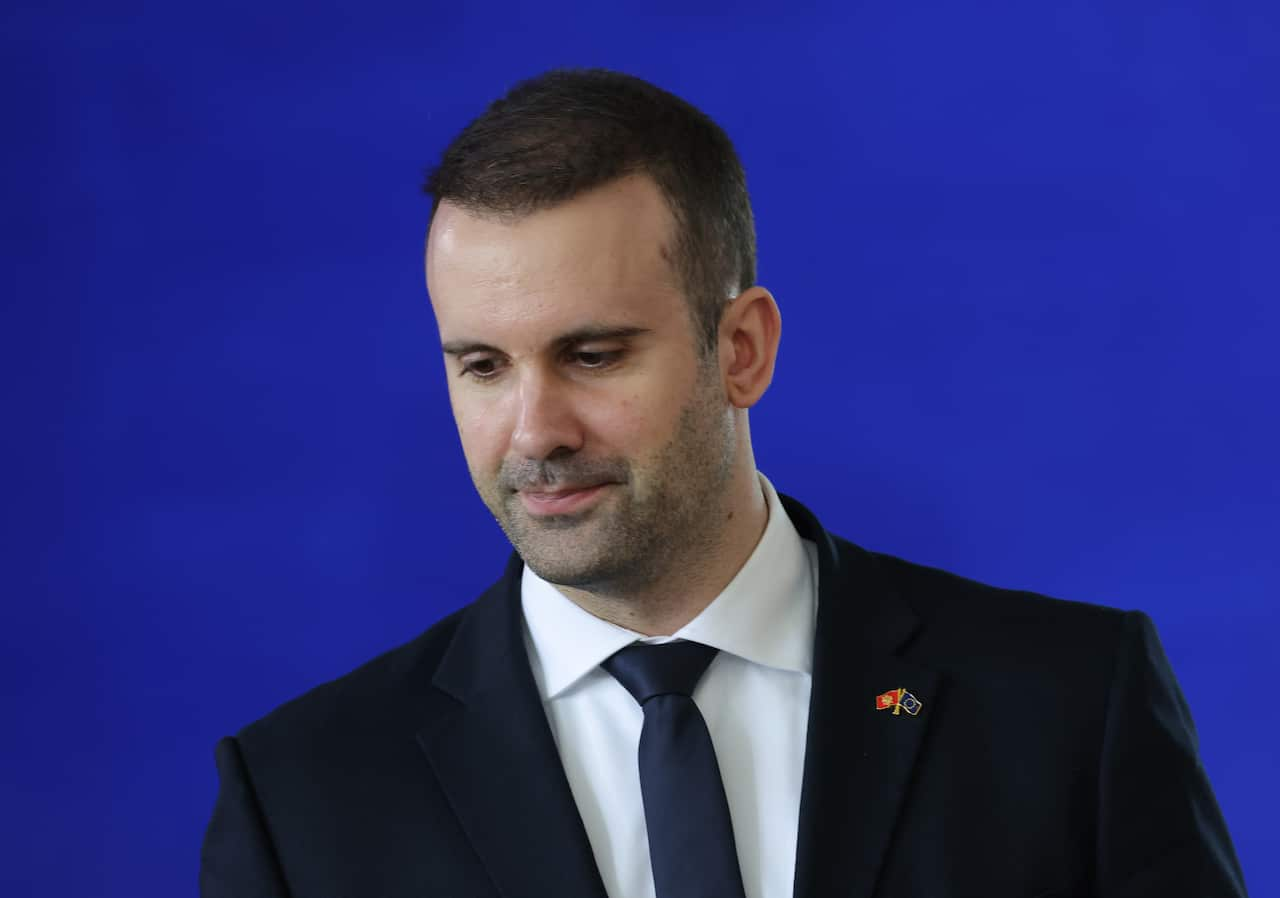 Milojko Spajic in a black suit, white shirt and blue tie, in front of a blue background.