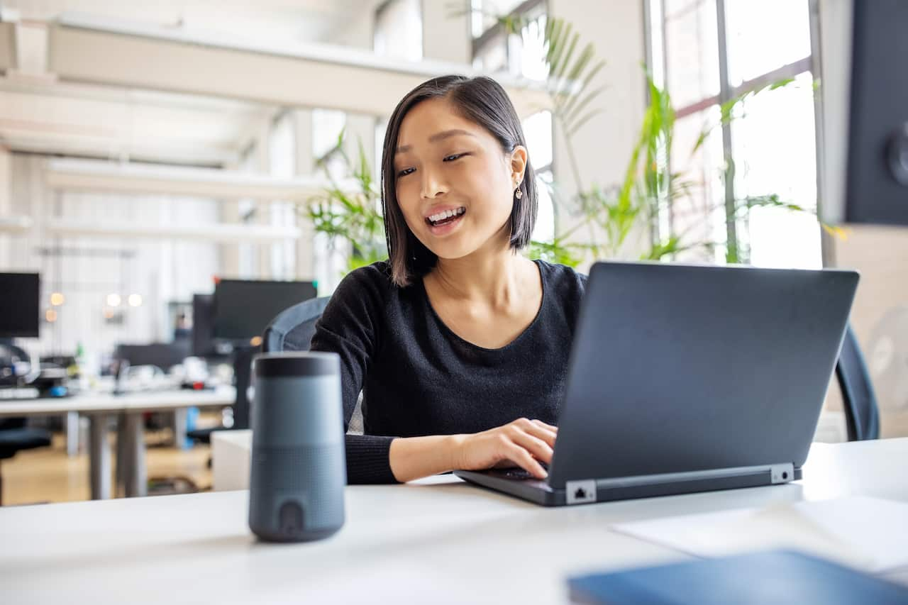 A woman using a virtual assistant at her desk while she uses a laptop.