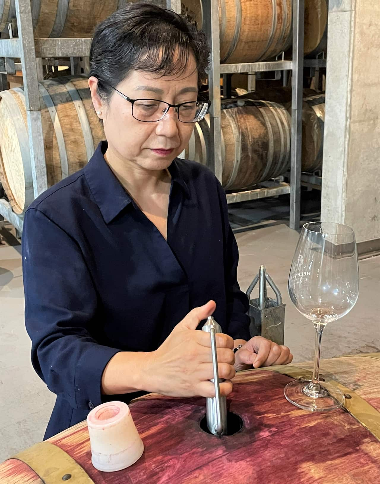 A woman in a blue shirt stands next to a barrel and draws wine for testing.