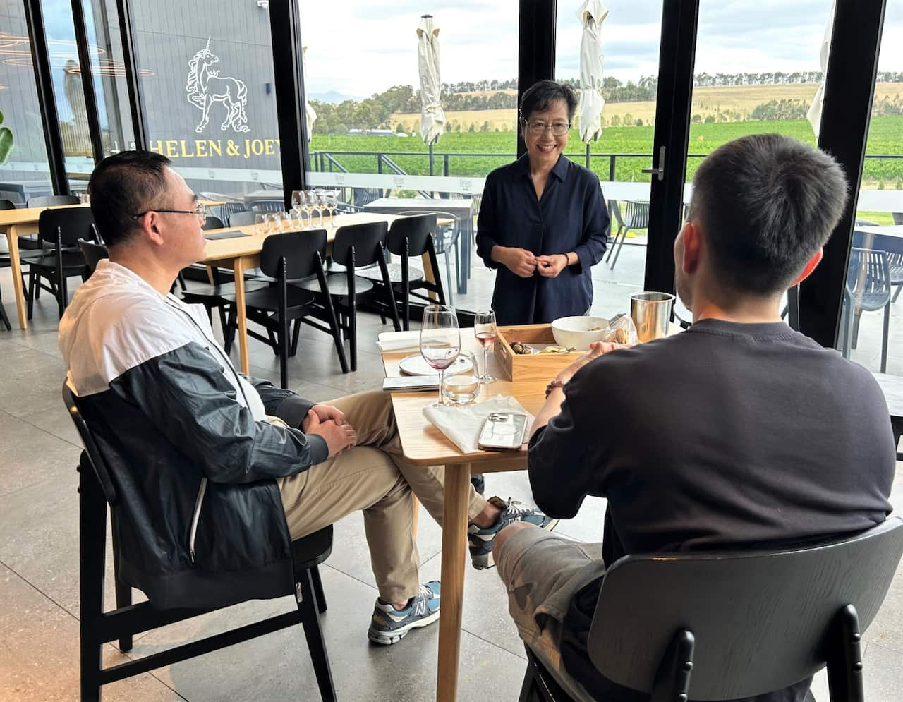 A woman in a blue shirt stands talking to two men seated at a winery.