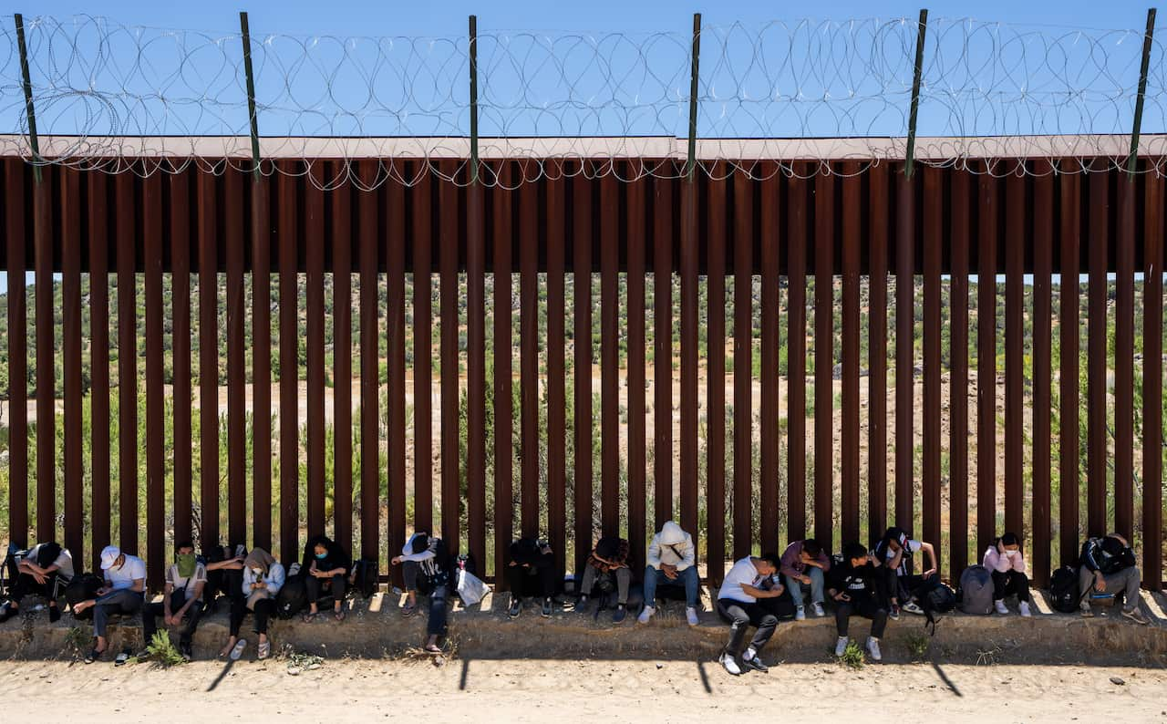 Migrants At The US-Mexico Border rest in the shade of the border wall.