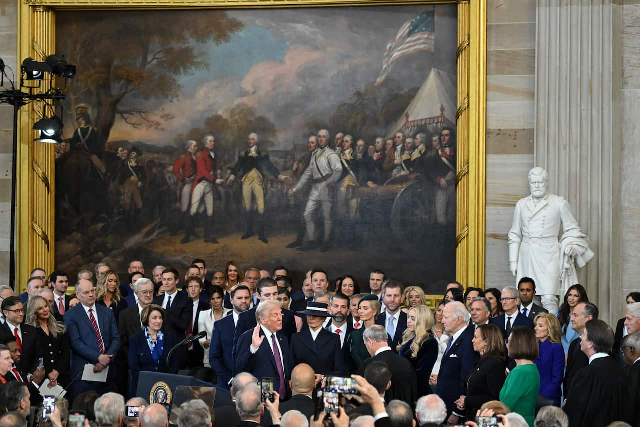 Man in crowded room taking oath, large paining in background.