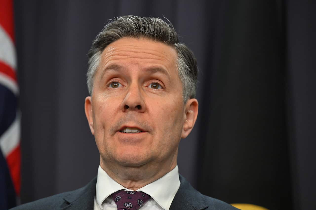 A man in a blue suit standing in front of a dark blue curtain and Australian flag.