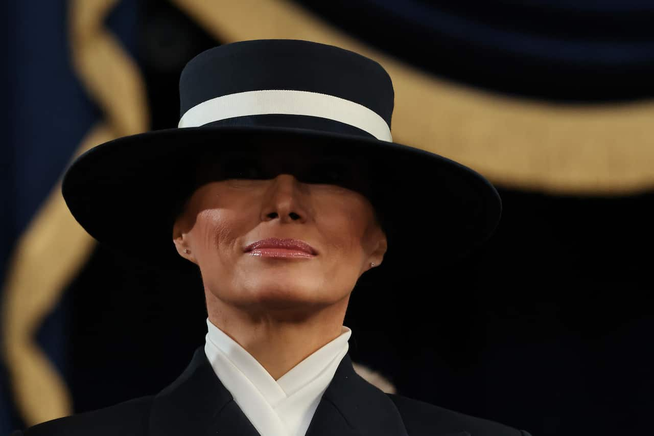 A woman in formal attire and a black hat with a white stripe obscures her eyes.