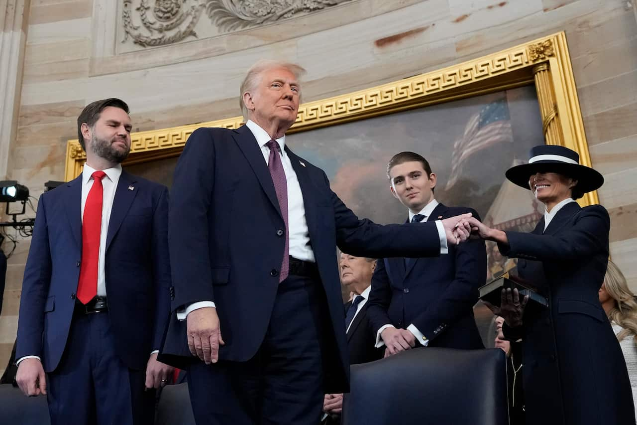 Donald Trump is holding the hand of a woman in a wide-brimmed hat as people look on and smile.