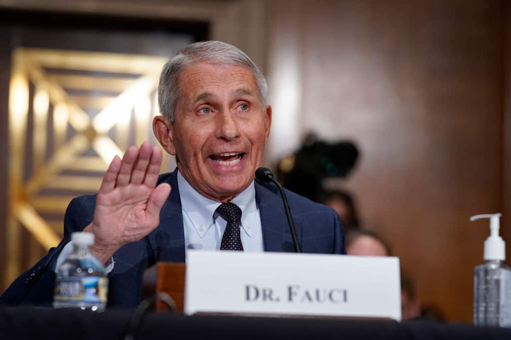 A man speaks at a podium with one hand raised, with a Dr. Fauci name tag placed in front of him on the podium.