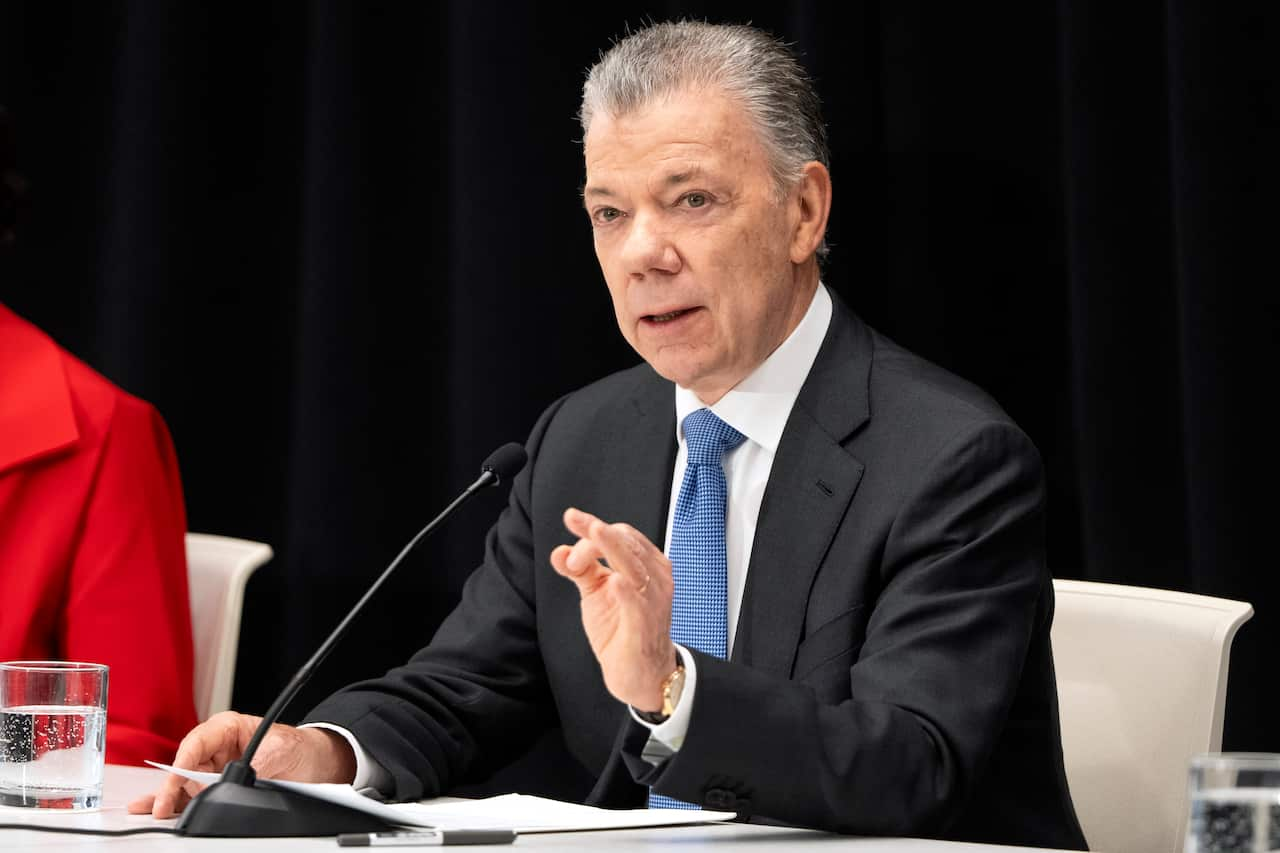 A man in a black suit, white shirt and blue tie sits at a desk and speaks into a microphone.