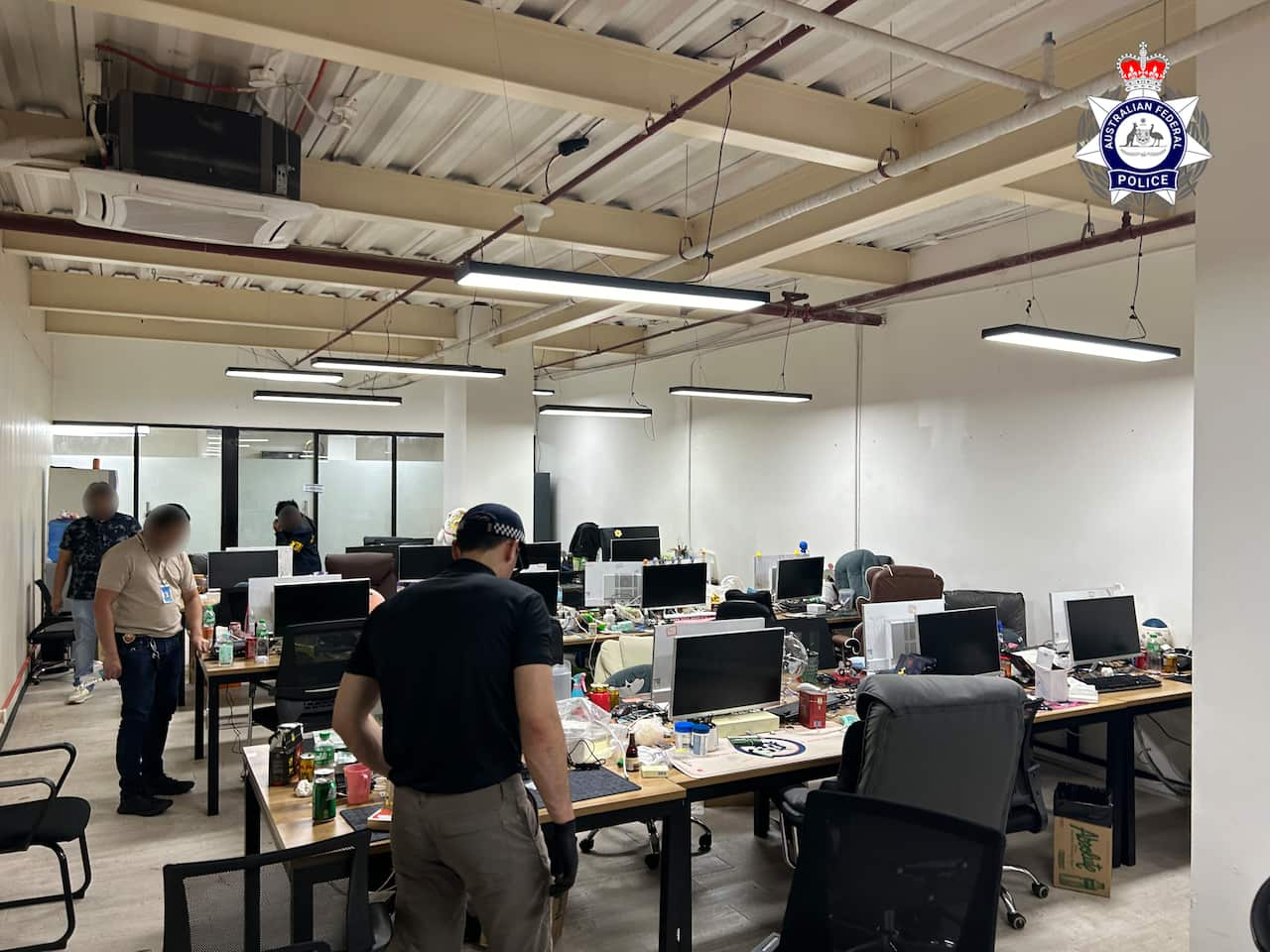 Police officers conduct a raid in an office space filled with rows of desks, computers, and chairs.