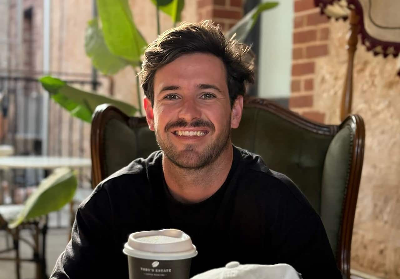 A man in a black T-shirt smiling while sitting in a cafe with a takeaway coffee on the table in front of him.