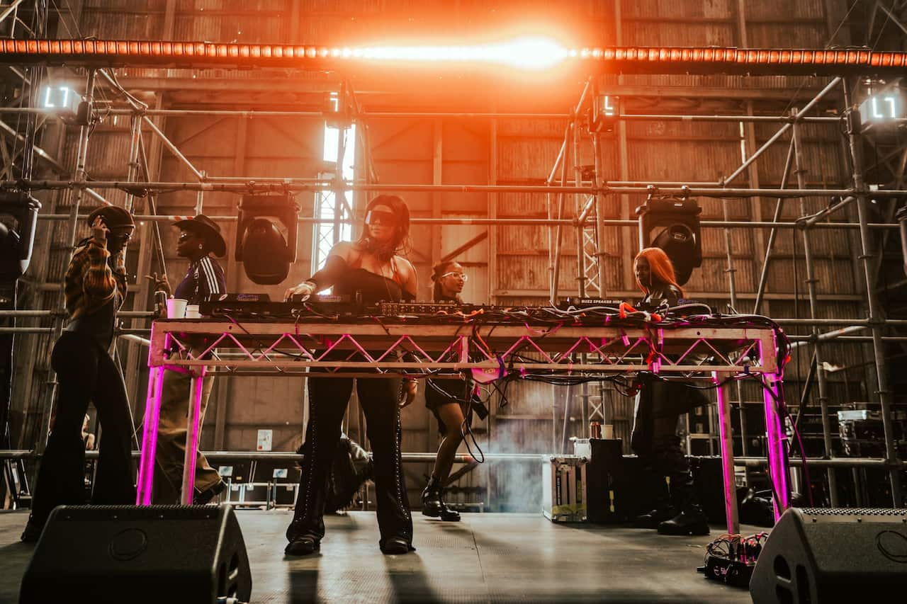 A DJ wearing sunglasses stands at her turntable on a stage in a warehouse-like room, with two people standing behind her