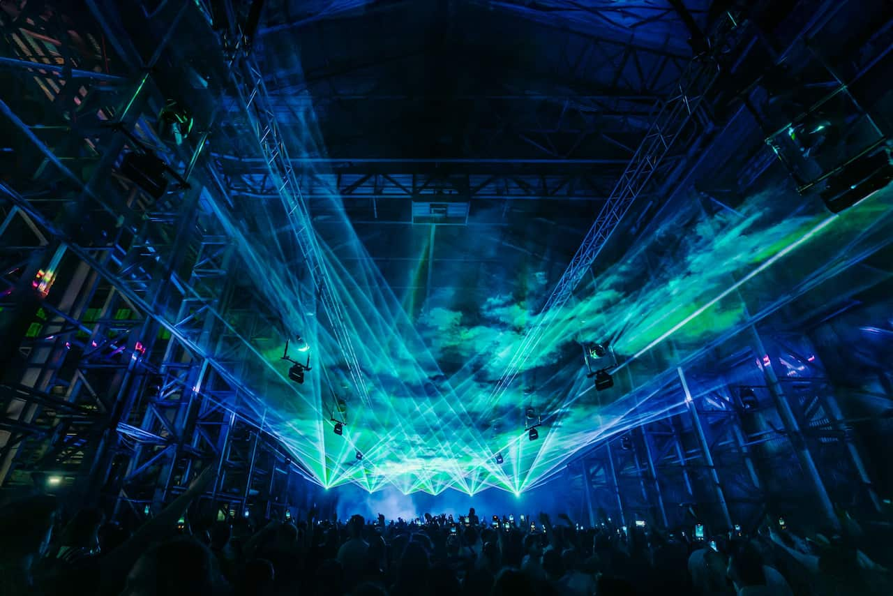 Blue and green lasers illuminate the ceiling of a large festival venue crowded with people 