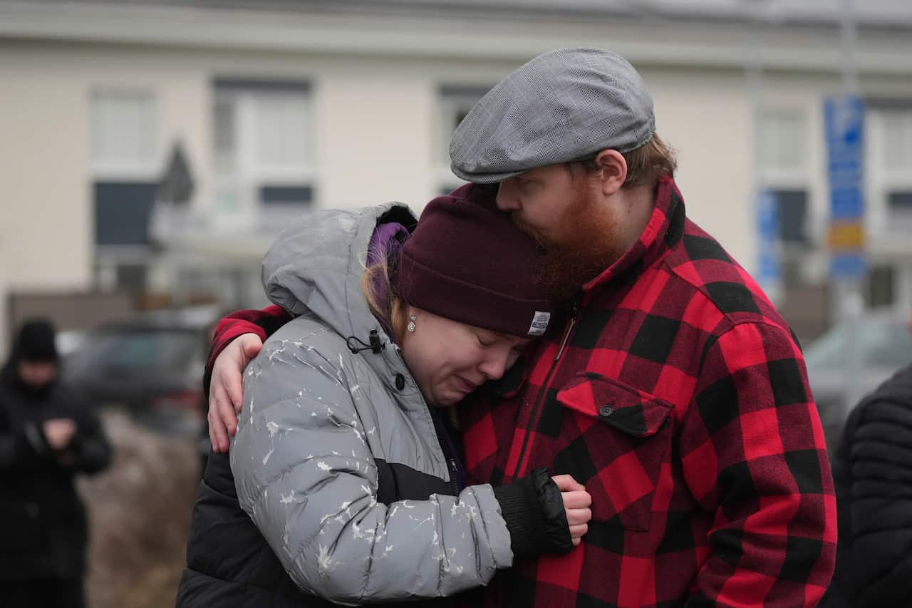 A man and woman embrace and outside. The woman, who is crying, is wearing a grey parka and black beanie and the man is wearing a black and red checked shirt and grey flat cap