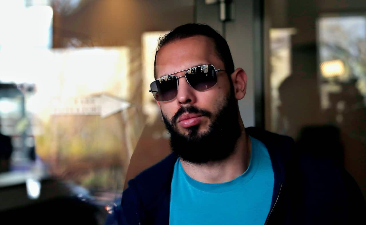 A man wearing a blue round-neck t-shirt, black pullover, and sunglasses stands outside a building.