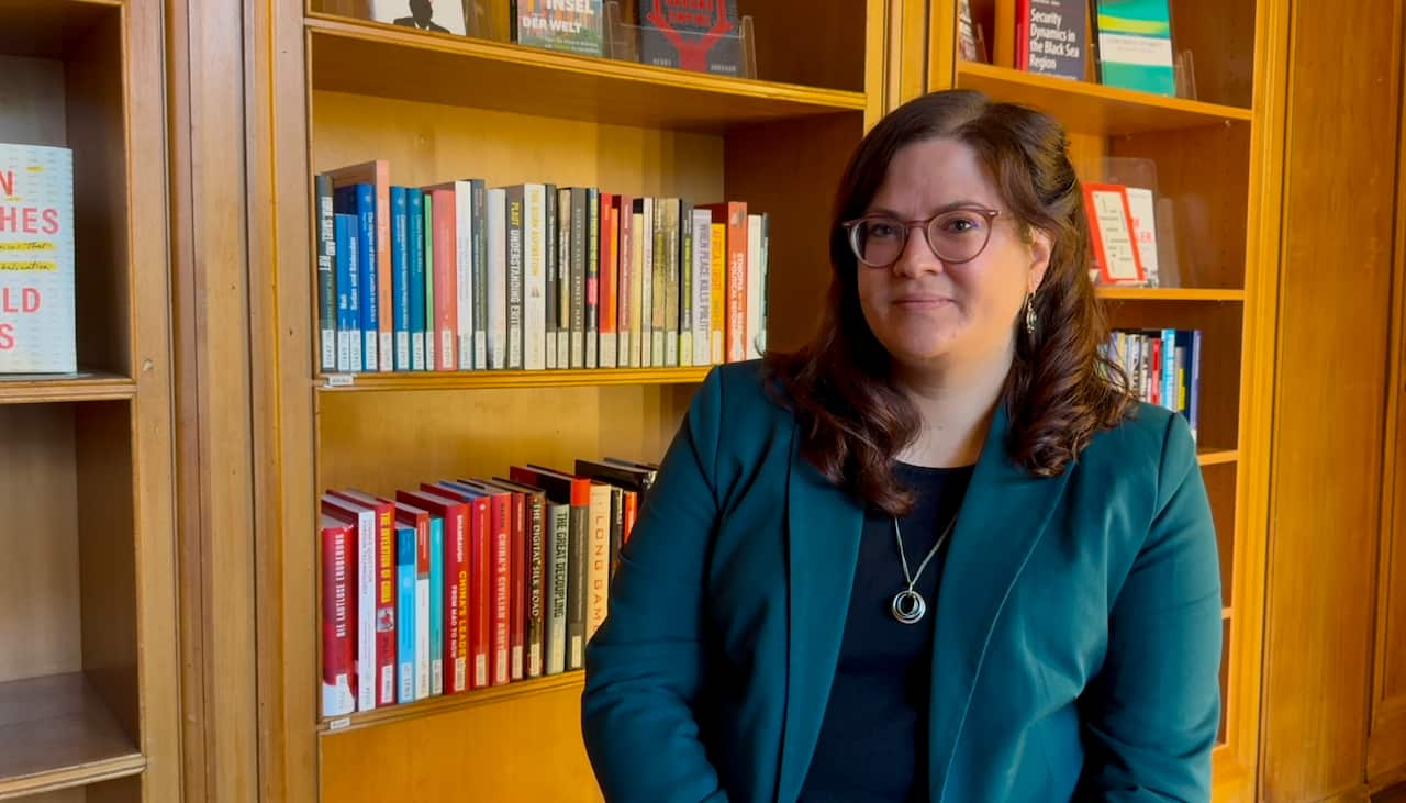 A woman is standing with books behind her. She has brown hair, glasses and is wearing a blue blazer.