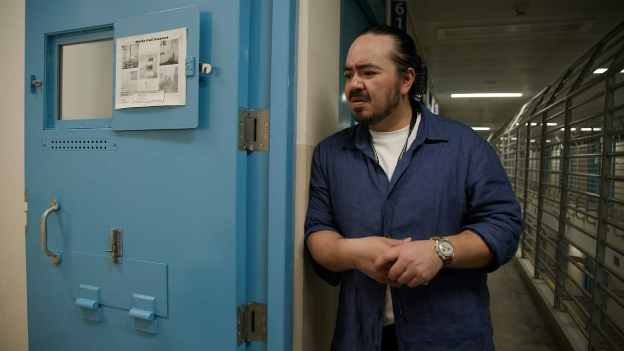 Adam Liaw in a blue over shirt looks concerned as he looks into a cell by a light blue cell door. 