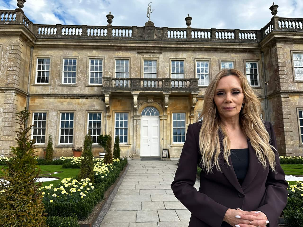 Karla, with long blonde hair and wearing a black suit, hands together, stands before a large stately home in sandstone. 