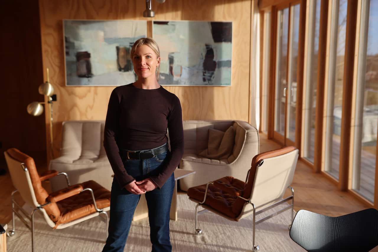  A woman in a black, long-sleeved shirt and blue jeans stands before stylish furniture in warm tones. 