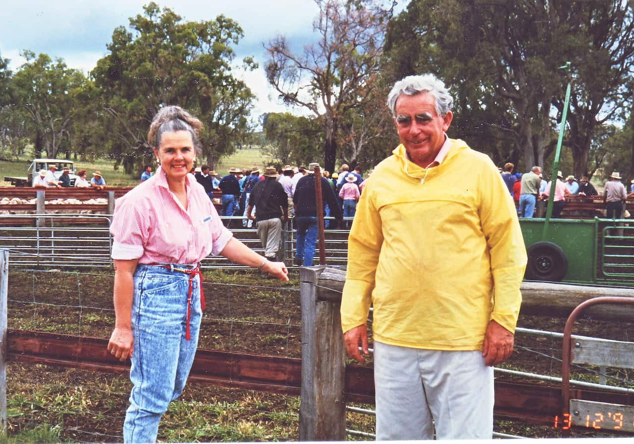 A man and a woman stand at some cattle yards, with a group of farmers in the background. The woman is wearing a pink shirt and blue jeans and the man is wearing a yellow rainjacket and light-coloured trousers 