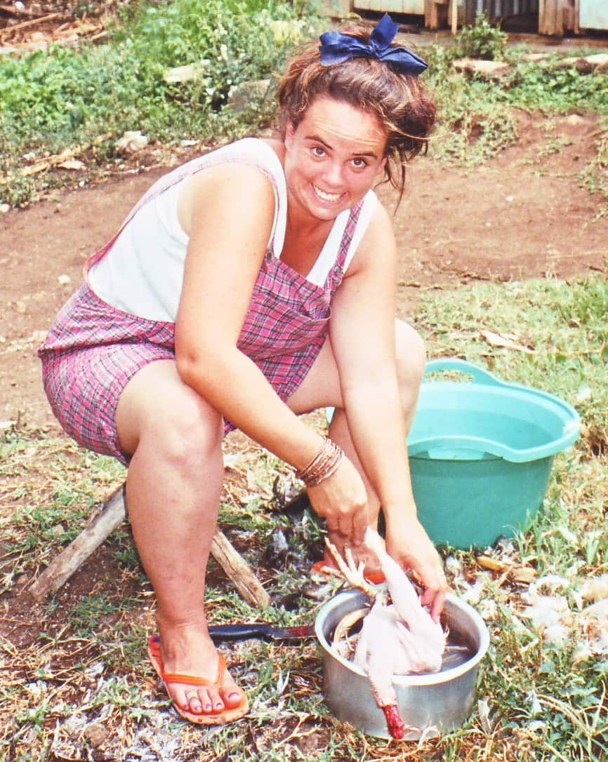 A young woman smiles as she squats and removes feathers from a chicken outside.