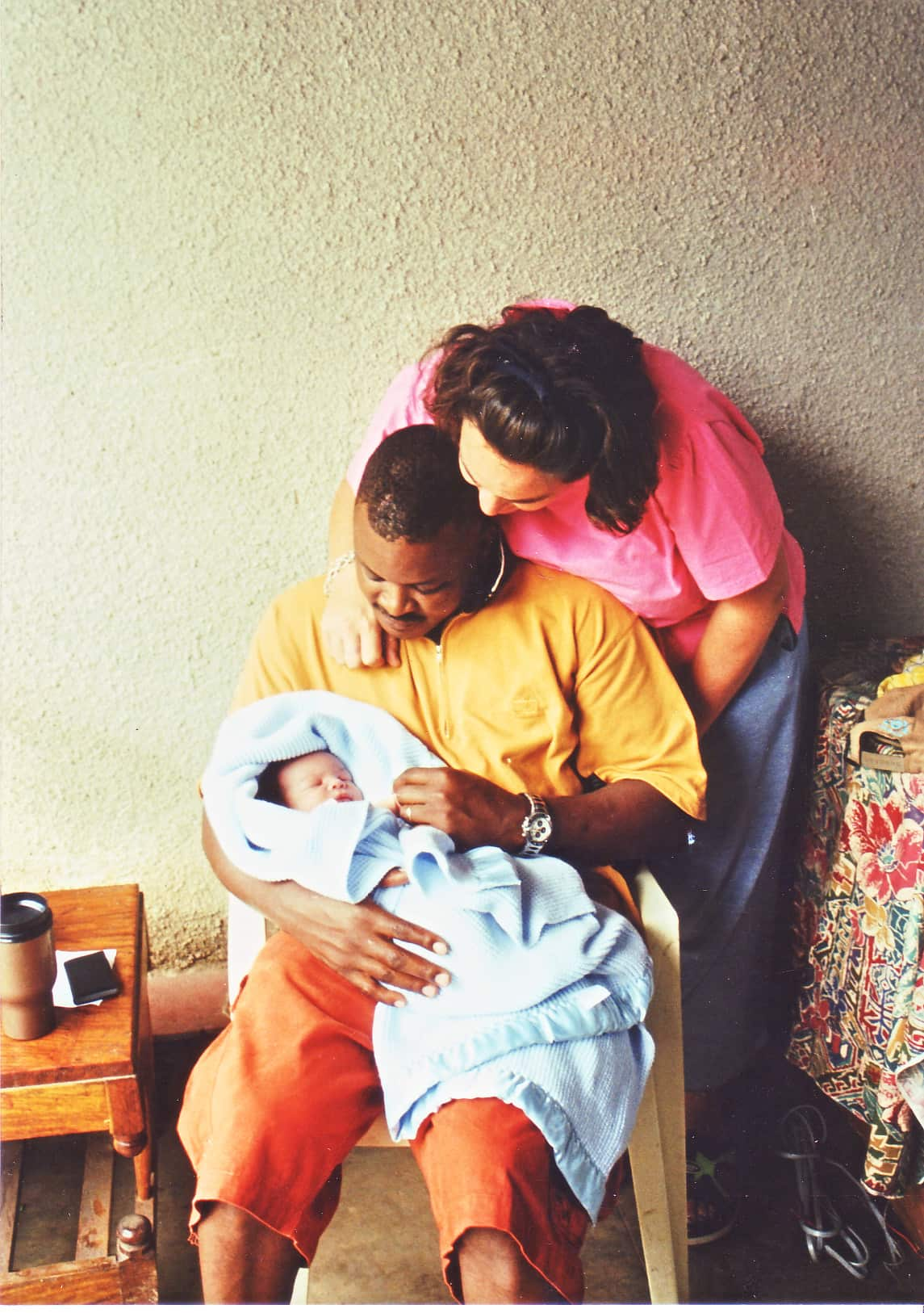 A man is sitting on a chair holding a newborn baby in a light-blue blanket. A woman stands behind him looking at the baby