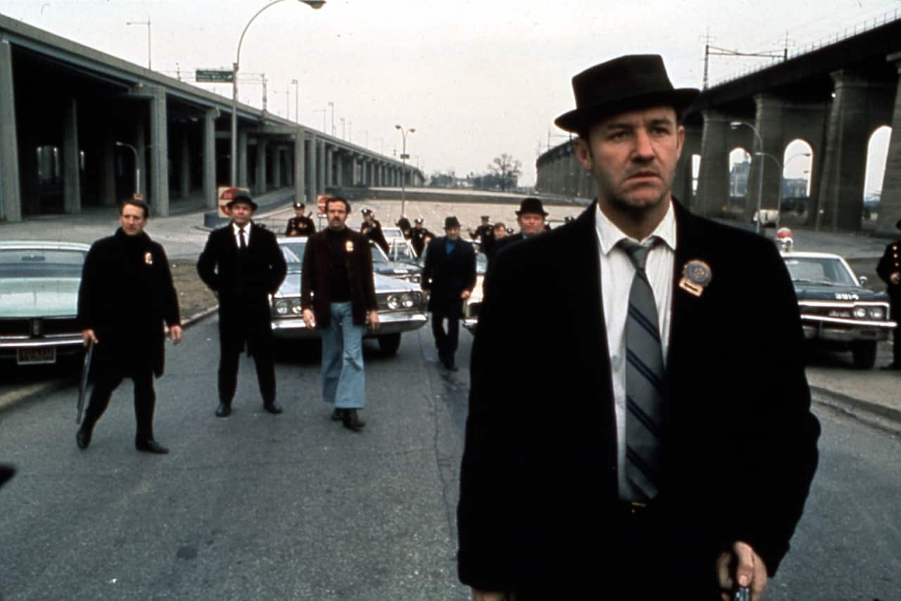 A young man in a black suit, matching hat, and police badge walks ahead of other officers on a wide road during a film shoot, with cars parked behind them.