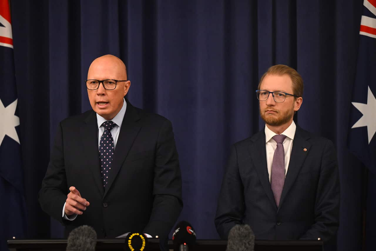 Two men in dark navy suits, look serious. The man on the left is points ahead while talking.