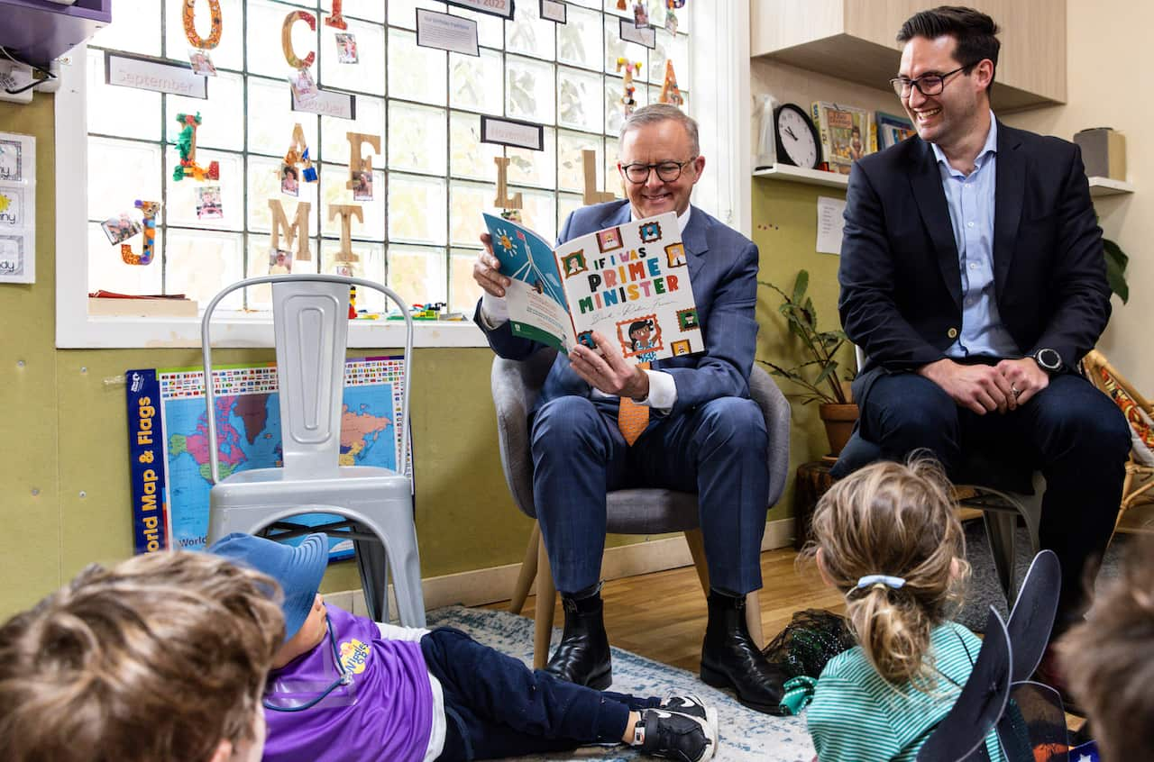 Anthony Albanese is sitting in a chair reading a book to children who are on the floor. Another man in a dark suit is sitting next to him.