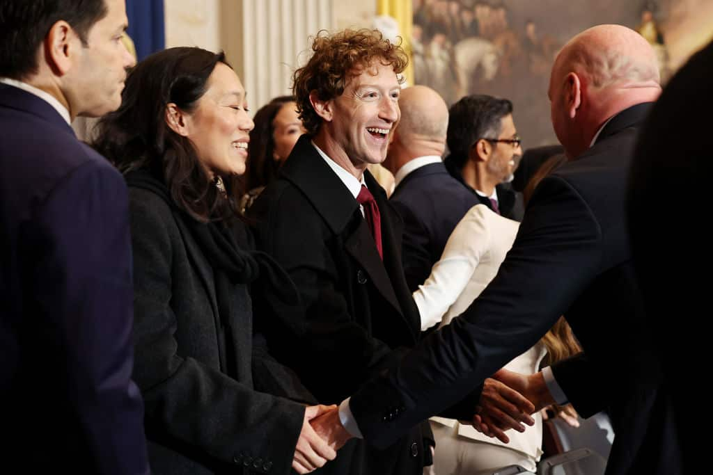 Mark Zuckerberg is laughing as he shakes hands with another man. 