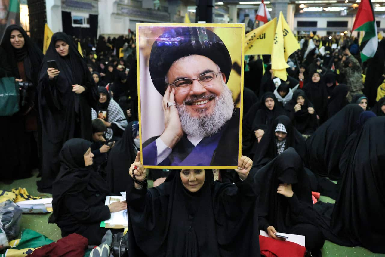 A woman holds up a man's portrait with a large crowd in black robes in the background.