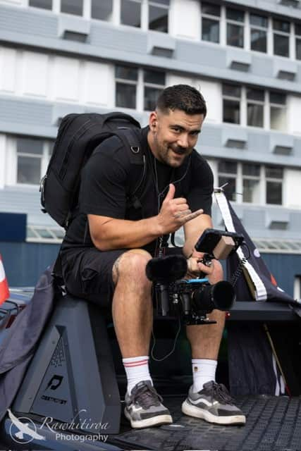 A man in a black T-shirt, shorts, and a backpack sits casually, holding a camera in one hand while making a 'hang loose' gesture with the other.